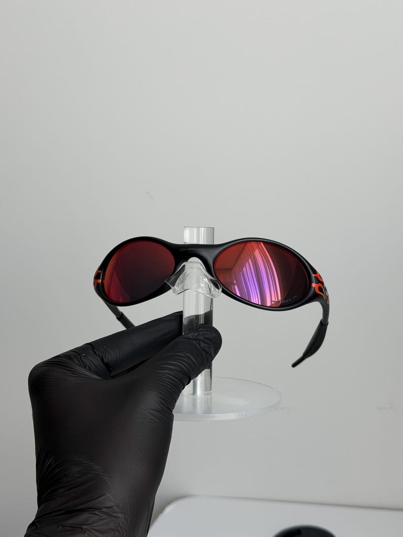 May include: Black sunglasses with red lenses and orange accents on the temples. The sunglasses are held on a clear display stand. The lenses reflect a purple hue.