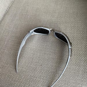 May include: Silver-framed sunglasses with dark lenses. The sunglasses have a sleek, modern design with curved temples. The lenses are rectangular and appear to be polarized. The sunglasses are resting on a textured, light gray surface.