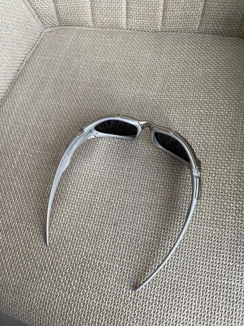 May include: Silver-framed sunglasses with dark lenses. The sunglasses have a sleek, modern design with curved temples. The lenses are rectangular and appear to be polarized. The sunglasses are resting on a textured, light gray surface.