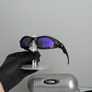 May include: Black sunglasses with purple lenses are displayed on a clear stand. The sunglasses have a sleek, modern design. A silver case and a white box with text are also visible.