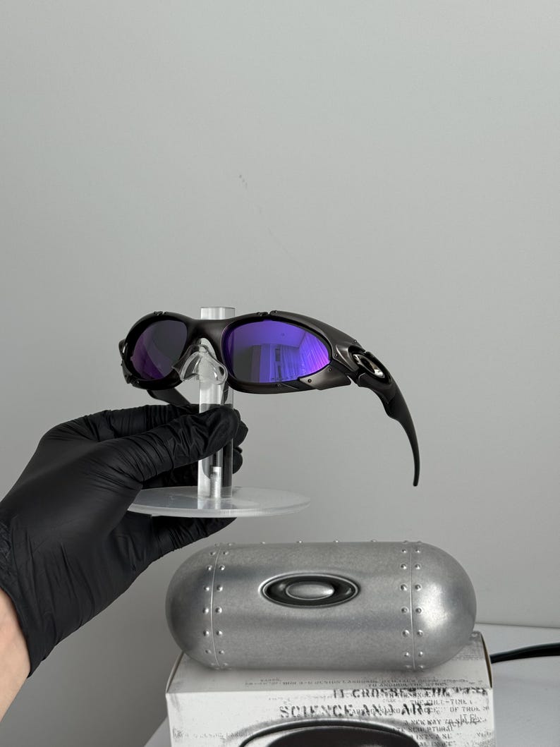 May include: Black sunglasses with purple lenses are displayed on a clear stand. The sunglasses have a sleek, modern design. A silver case and a white box with text are also visible.