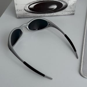 Pu&ograve; includere: Occhiali da sole con montatura argentata e lenti scure, dal design elegante e moderno. Le aste hanno impugnature in gomma nera per il comfort. Gli occhiali da sole sono esposti con la loro scatola originale, con la scritta "SCIENCE AND ART".