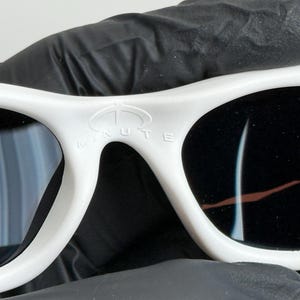 May include: Close-up of white sunglasses with black lenses. The sunglasses have a curved frame and the word "MINUTE" is printed on the top of the frame. The sunglasses are held by a black gloved hand.