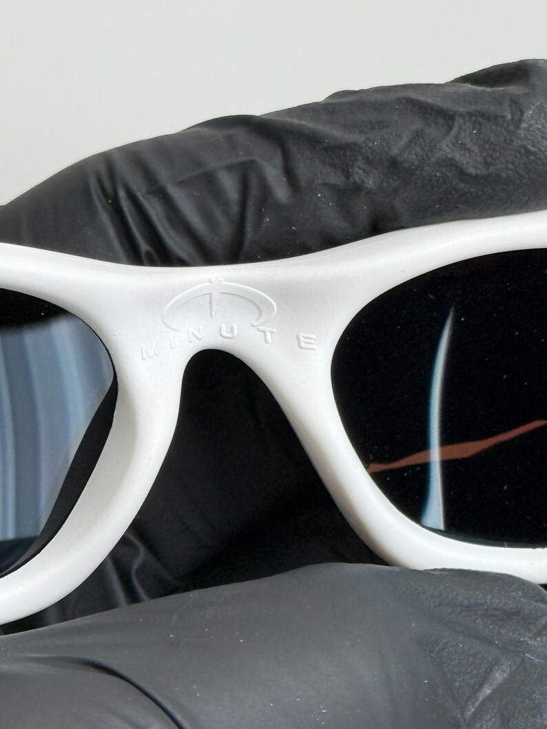 May include: Close-up of white sunglasses with black lenses. The sunglasses have a curved frame and the word "MINUTE" is printed on the top of the frame. The sunglasses are held by a black gloved hand.