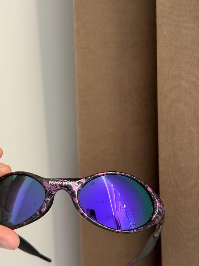 May include: Oval-shaped sunglasses with a black and pink speckled frame and blue mirrored lenses. The text "BRAIN DEAD" is printed on the top of the frame. The sunglasses have black arms.