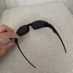 May include: Black sunglasses with a rectangular frame and dark lenses. The sunglasses have a sleek, modern design with a curved temple. The sunglasses are resting on a neutral-coloured surface.