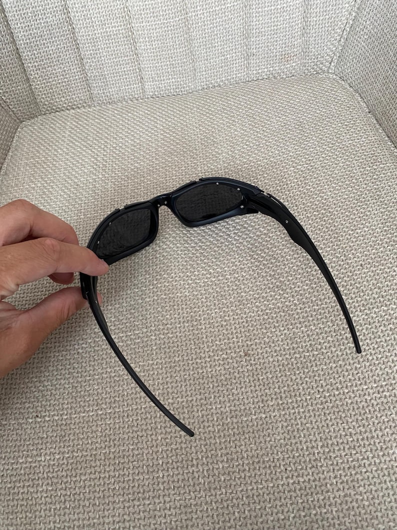 May include: Black sunglasses with a rectangular frame and dark lenses. The sunglasses have a sleek, modern design with a curved temple. The sunglasses are resting on a neutral-coloured surface.