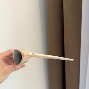 May include: Tan-colored sunglasses with dark lenses. The sunglasses have a sleek, modern design with a curved frame and straight arms. The arms are a lighter shade of tan with a silver accent at the tips. The brand name is visible on the arm.