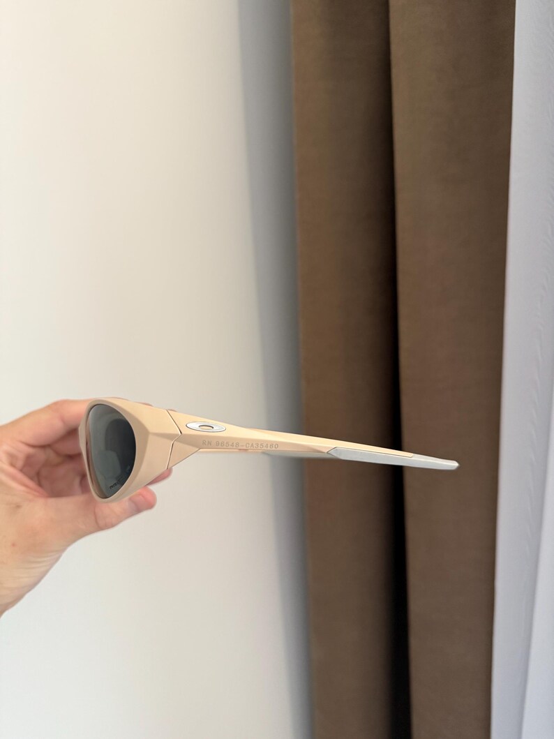 May include: Tan-colored sunglasses with dark lenses. The sunglasses have a sleek, modern design with a curved frame and straight arms. The arms are a lighter shade of tan with a silver accent at the tips. The brand name is visible on the arm.