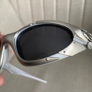 May include: Silver-framed sunglasses with dark lenses. The sunglasses have a sleek, modern design with a riveted metal frame and white arms. The lenses are polarized, as indicated by the text on the lens.