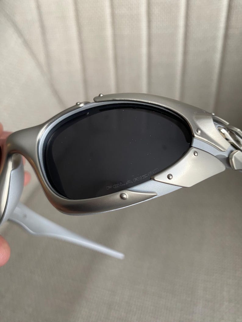 May include: Silver-framed sunglasses with dark lenses. The sunglasses have a sleek, modern design with a riveted metal frame and white arms. The lenses are polarized, as indicated by the text on the lens.