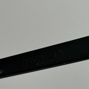 May include: A close-up of a black object with the word "Frogskins" embossed on it. The object is set against a plain, light gray background. The text is in a sans-serif font.