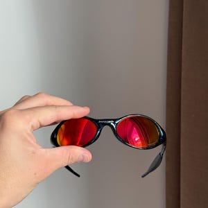 Puede incluir: Gafas de sol negras con lentes de espejo rojas. Las gafas de sol son sostenidas por una mano. La montura curva tiene una peque&ntilde;a marca impresa en la parte superior. Las lentes reflejan el entorno.