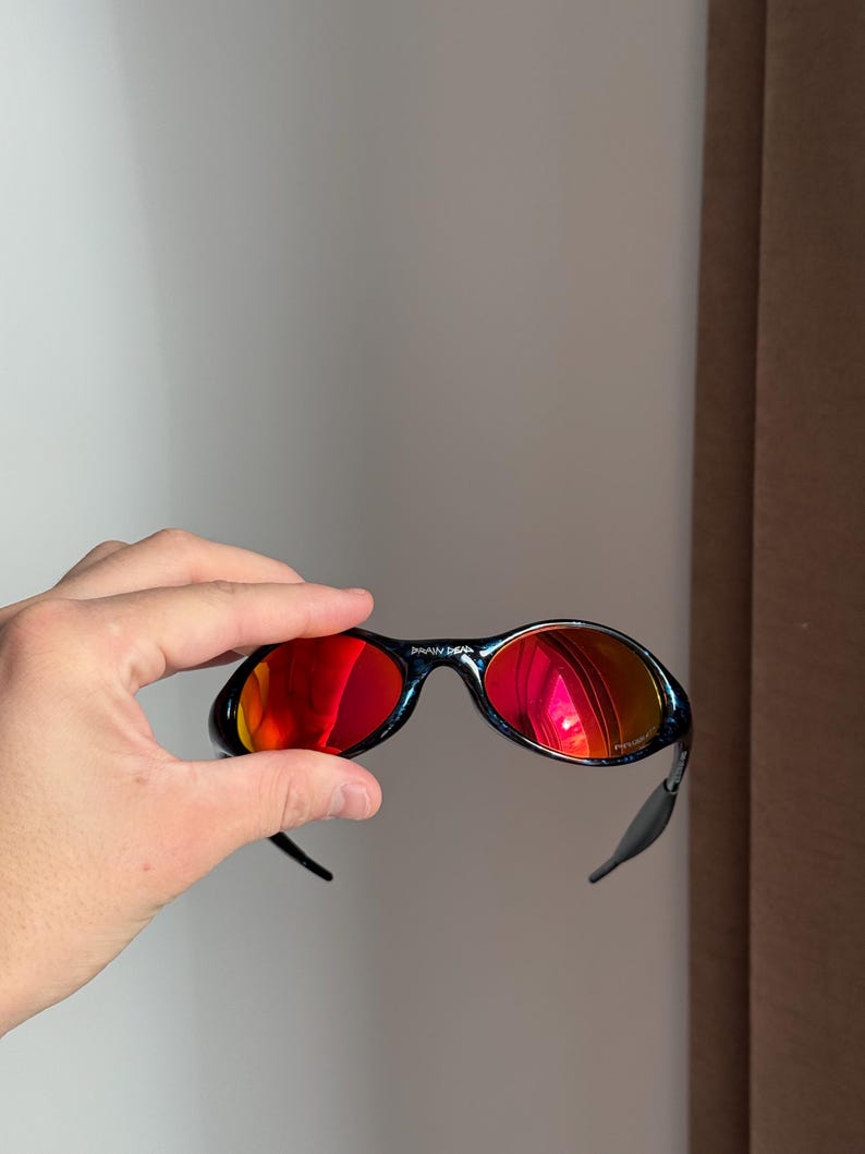 Puede incluir: Gafas de sol negras con lentes de espejo rojas. Las gafas de sol son sostenidas por una mano. La montura curva tiene una peque&ntilde;a marca impresa en la parte superior. Las lentes reflejan el entorno.