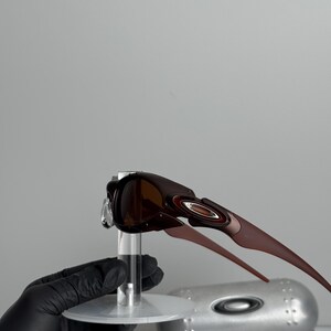 May include: Brown sunglasses with a dark frame and brown lenses. The sunglasses have a sleek, modern design with a curved shape and a metallic accent on the temples. The sunglasses are displayed on a clear stand.