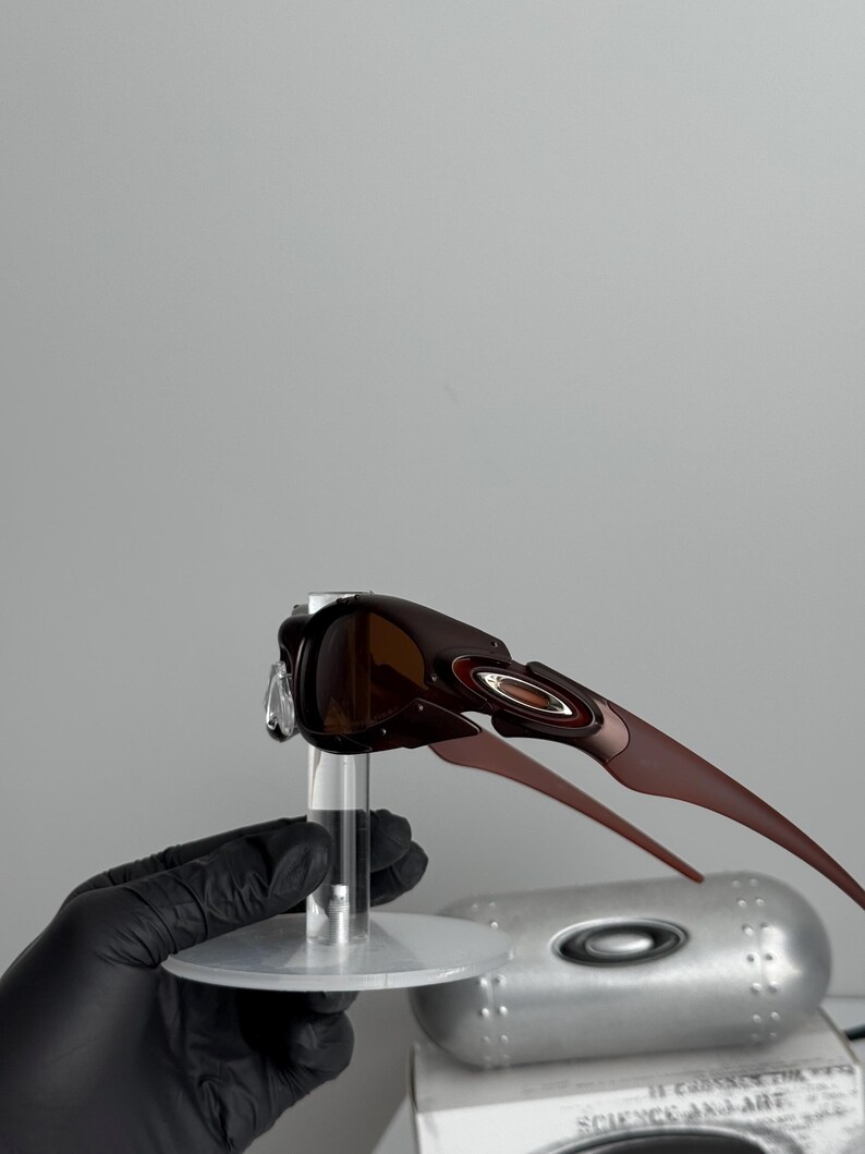May include: Brown sunglasses with a dark frame and brown lenses. The sunglasses have a sleek, modern design with a curved shape and a metallic accent on the temples. The sunglasses are displayed on a clear stand.