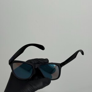 May include: Black sunglasses with blue-tinted lenses. The sunglasses have a rectangular frame and curved temples. The brand name "Oakley" is visible on the left temple. The sunglasses are held by a gloved hand.