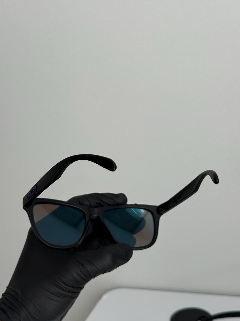 May include: Black sunglasses with blue-tinted lenses. The sunglasses have a rectangular frame and curved temples. The brand name "Oakley" is visible on the left temple. The sunglasses are held by a gloved hand.