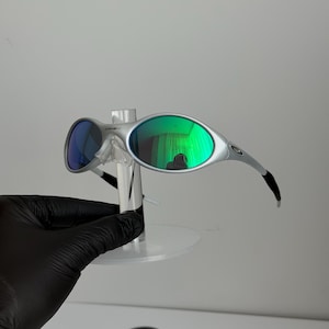 May include: Silver sunglasses with green and blue lenses. The sunglasses have a modern design with curved arms and a small logo on the frame. The lenses reflect a green hue, and the sunglasses are displayed on a clear stand.