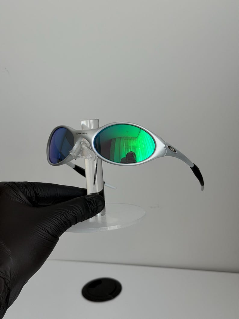May include: Silver sunglasses with green and blue lenses. The sunglasses have a modern design with curved arms and a small logo on the frame. The lenses reflect a green hue, and the sunglasses are displayed on a clear stand.