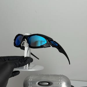 May include: Black sunglasses with blue mirrored lenses are displayed on a clear stand. The sunglasses have a sleek, sporty design with curved temples. A silver case with the brand logo is visible below the sunglasses.