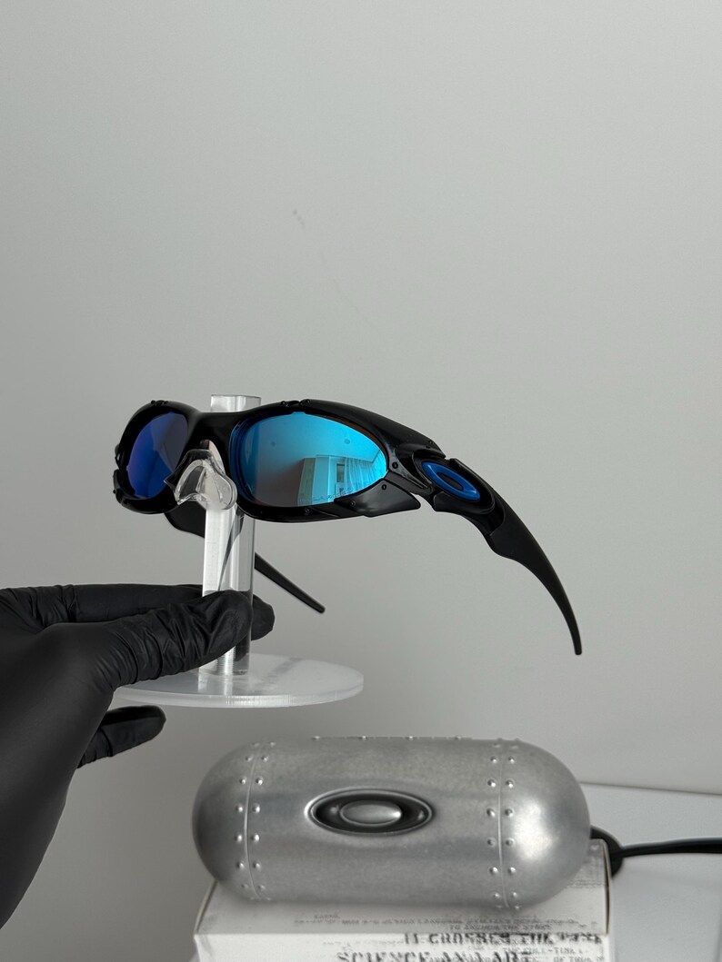 May include: Black sunglasses with blue mirrored lenses are displayed on a clear stand. The sunglasses have a sleek, sporty design with curved temples. A silver case with the brand logo is visible below the sunglasses.