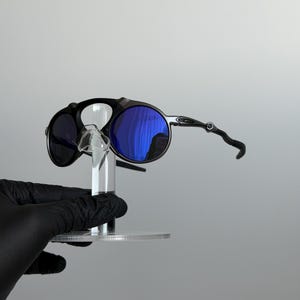 May include: Sunglasses featuring a matte black frame with circular lenses and a vibrant blue reflective coating. The design includes a distinct bridge structure connecting the two round rims, paired with slender, contoured temples.