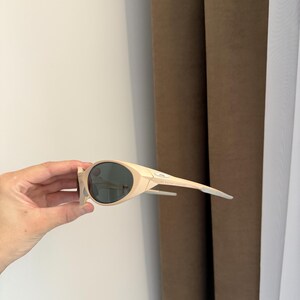 May include: Cream-colored sunglasses with dark lenses. The sunglasses have a sleek, modern design with a curved frame and straight temples. The lenses are a dark gray color, and the frame is a light beige. The sunglasses are held by a hand.