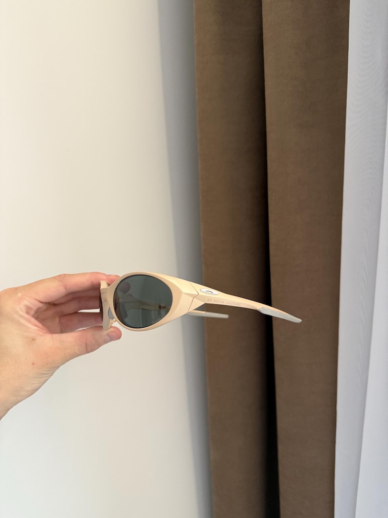 May include: Cream-colored sunglasses with dark lenses. The sunglasses have a sleek, modern design with a curved frame and straight temples. The lenses are a dark gray color, and the frame is a light beige. The sunglasses are held by a hand.