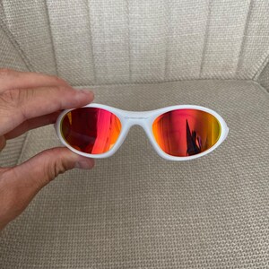 Puede incluir: Gafas de sol blancas con lentes reflectantes rojas y naranjas. Las gafas tienen una montura blanca y un dise&ntilde;o deportivo. El logotipo de Oakley es visible en la montura. Las gafas son sostenidas por una mano.