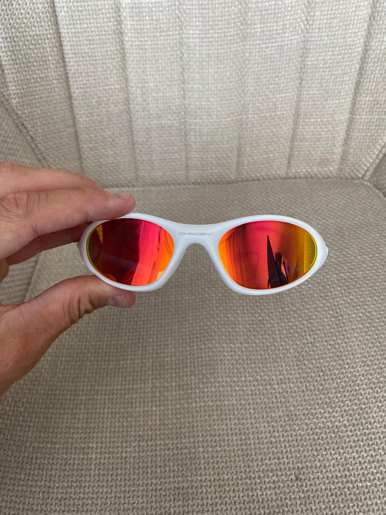 Puede incluir: Gafas de sol blancas con lentes reflectantes rojas y naranjas. Las gafas tienen una montura blanca y un dise&ntilde;o deportivo. El logotipo de Oakley es visible en la montura. Las gafas son sostenidas por una mano.