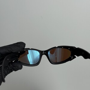 May include: Black sunglasses with a curved frame and tinted lenses. The lenses reflect blue and orange hues. The sunglasses have a sporty design.