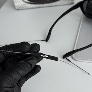 K&ouml;nnte beinhalten: Schwarze Sonnenbrille mit schlankem Design. Die B&uuml;gel sind d&uuml;nn und metallisch, mit schwarzen Ohrenteilen. Das Oakley-Logo ist auf dem B&uuml;gel sichtbar. Auf der Schachtel im Hintergrund steht "SCIENCE AND ART".