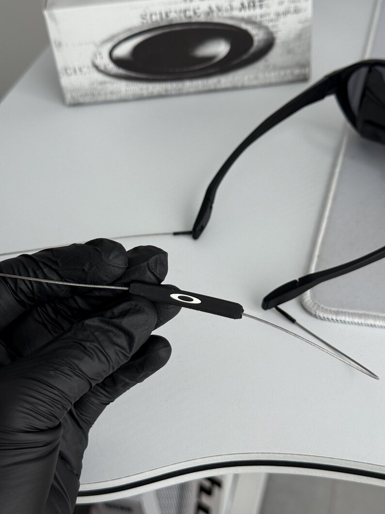 K&ouml;nnte beinhalten: Schwarze Sonnenbrille mit schlankem Design. Die B&uuml;gel sind d&uuml;nn und metallisch, mit schwarzen Ohrenteilen. Das Oakley-Logo ist auf dem B&uuml;gel sichtbar. Auf der Schachtel im Hintergrund steht "SCIENCE AND ART".