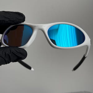 May include: White sunglasses with blue mirrored lenses. The sunglasses have a white frame with black accents on the temples. The Oakley logo is visible on the top of the frame.