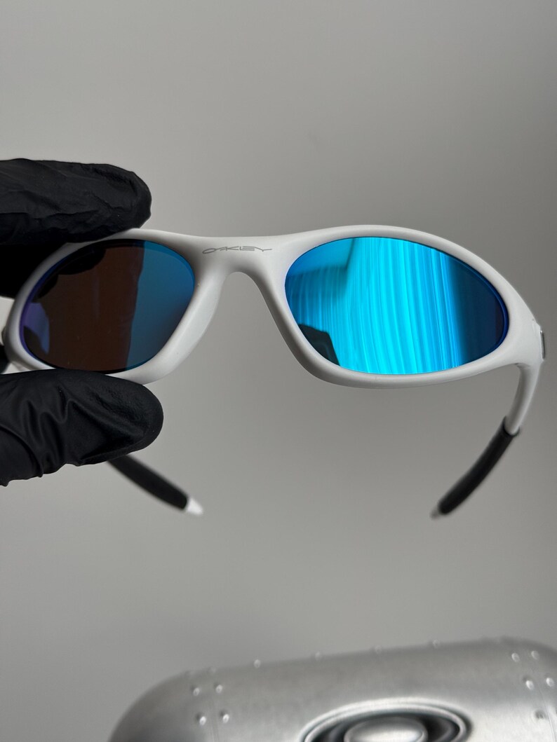 May include: White sunglasses with blue mirrored lenses. The sunglasses have a white frame with black accents on the temples. The Oakley logo is visible on the top of the frame.