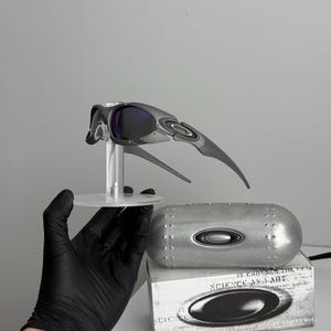 May include: Gray and black sports sunglasses with purple lenses, displayed on a clear stand. Includes a silver case and a box with the text "SCIENCE AND ART".