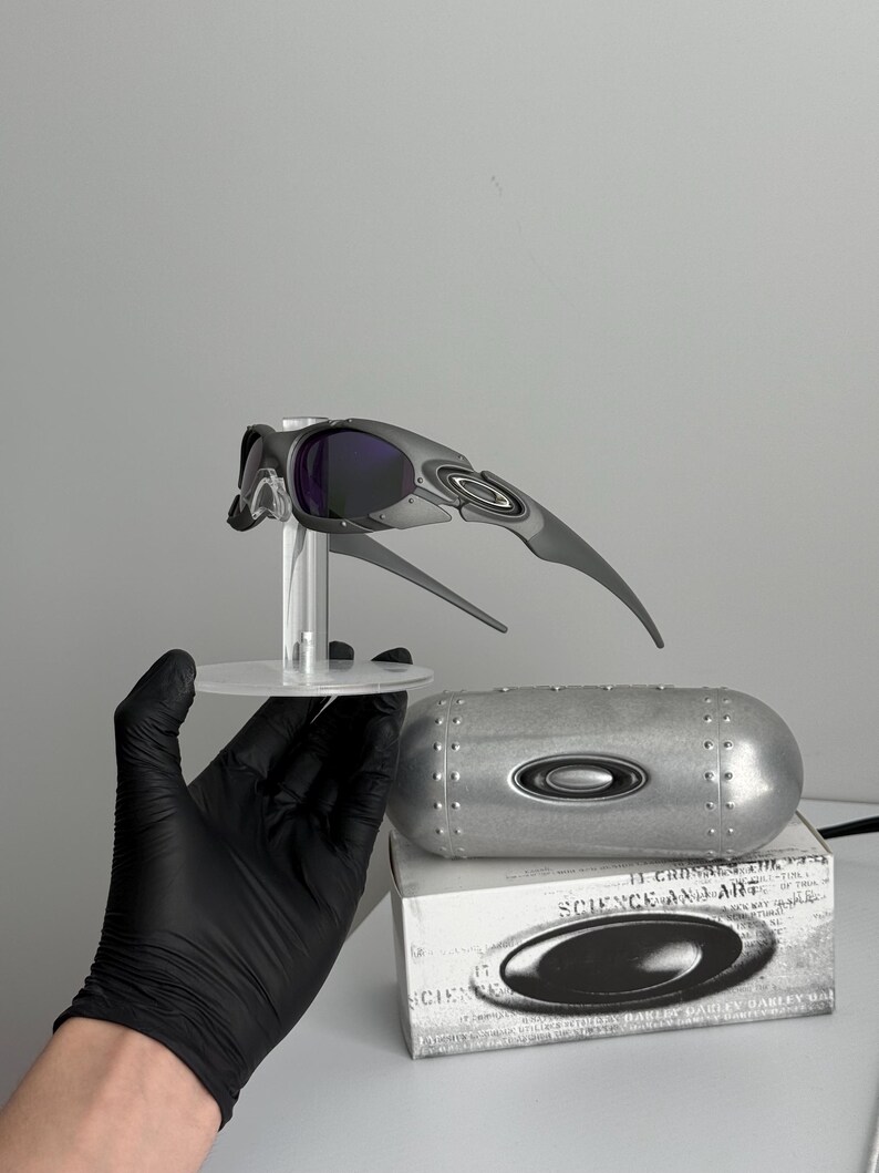 May include: Gray and black sports sunglasses with purple lenses, displayed on a clear stand. Includes a silver case and a box with the text "SCIENCE AND ART".