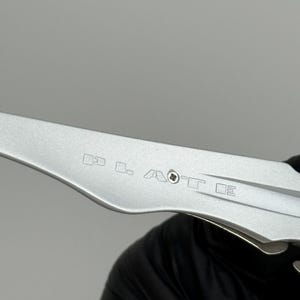 May include: A close-up of a silver metal tool with etched markings. The tool has a curved blade and a series of small, geometric symbols etched into the metal. The tool is held by a person wearing a black glove.