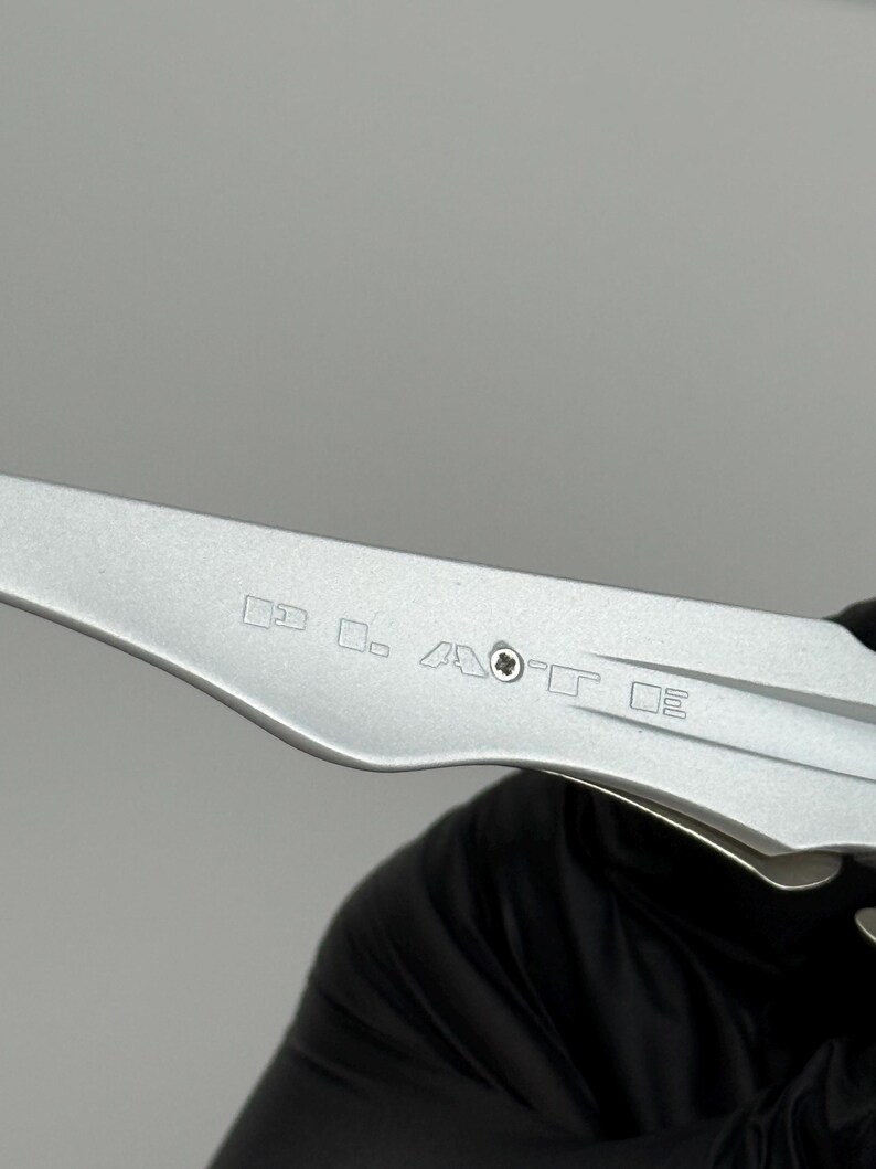 May include: A close-up of a silver metal tool with etched markings. The tool has a curved blade and a series of small, geometric symbols etched into the metal. The tool is held by a person wearing a black glove.