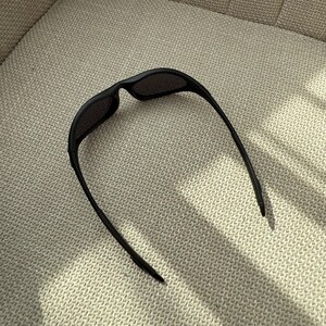 May include: Black sunglasses with a curved frame and dark lenses. The sunglasses are resting on a textured, light-coloured surface. The arms of the sunglasses extend outwards.