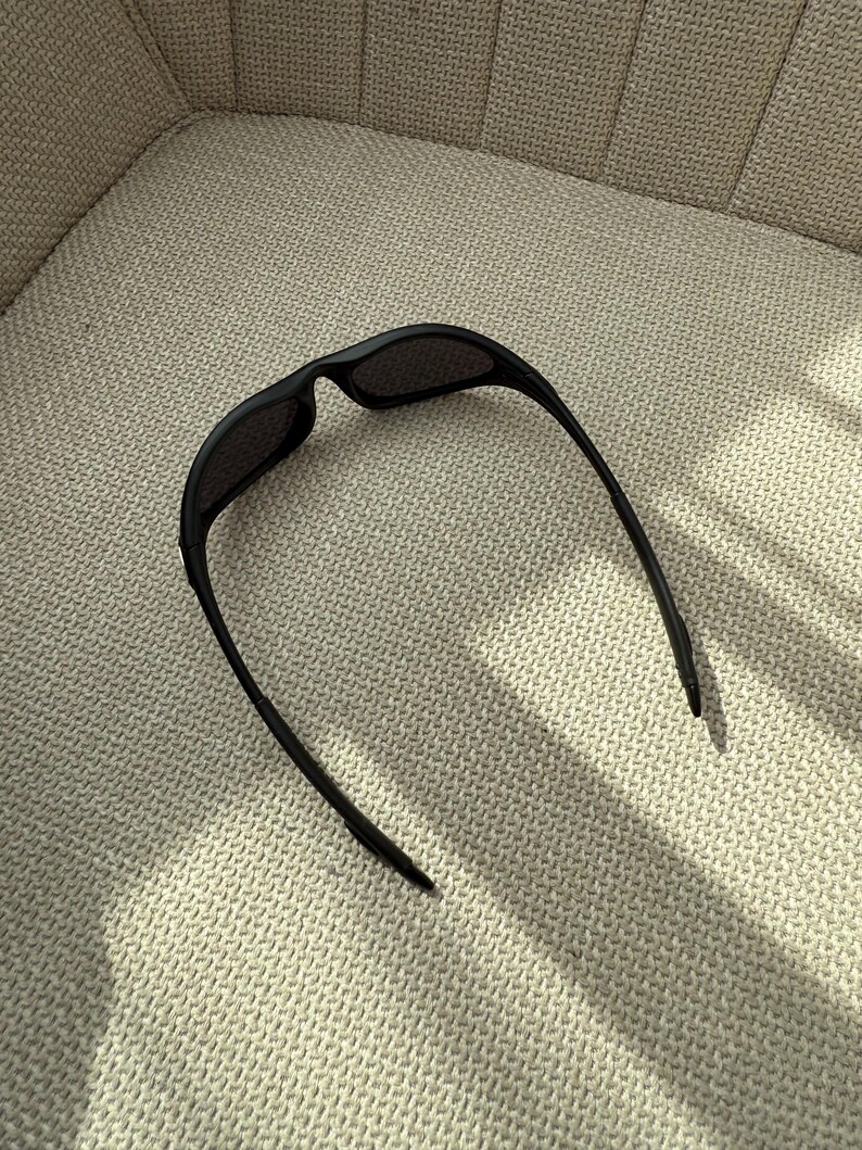 May include: Black sunglasses with a curved frame and dark lenses. The sunglasses are resting on a textured, light-coloured surface. The arms of the sunglasses extend outwards.