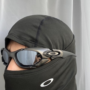 Può includere: Occhiali da sole grigio scuro con montatura argentata e lenti a specchio. Gli occhiali da sole sono indossati sopra un passamontagna nero. Il logo Oakley è visibile sul passamontagna.