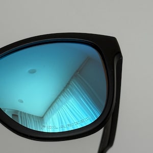 May include: Black sunglasses with blue polarized lenses. The lenses reflect a light blue hue, and the text "POLARIZED" is printed on the lens. The frame is a matte black.