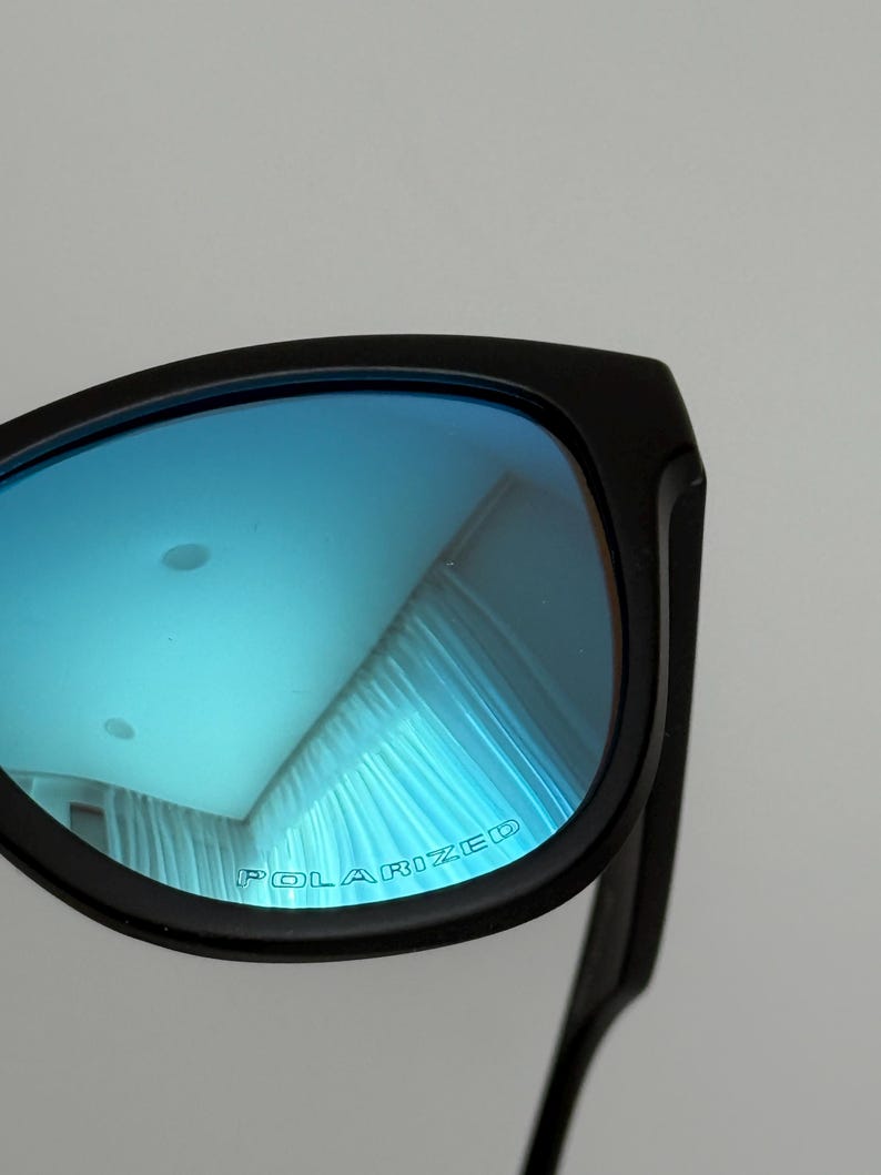 May include: Black sunglasses with blue polarized lenses. The lenses reflect a light blue hue, and the text "POLARIZED" is printed on the lens. The frame is a matte black.
