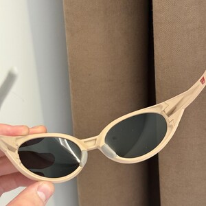 May include: Beige-colored sunglasses with dark lenses. The sunglasses have a sleek, curved frame and a small, rectangular detail on the temple. The lenses are oval-shaped and appear to be tinted. The sunglasses are held by a hand.