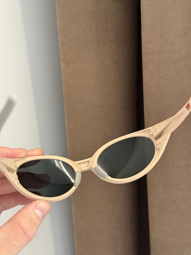 May include: Beige-colored sunglasses with dark lenses. The sunglasses have a sleek, curved frame and a small, rectangular detail on the temple. The lenses are oval-shaped and appear to be tinted. The sunglasses are held by a hand.