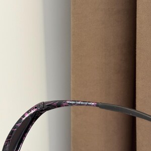 May include: Black sunglasses with a purple and black patterned frame. The glasses have a curved design and a dark lens. The text "008450-1259 59□22 135 3N" is printed on the frame.