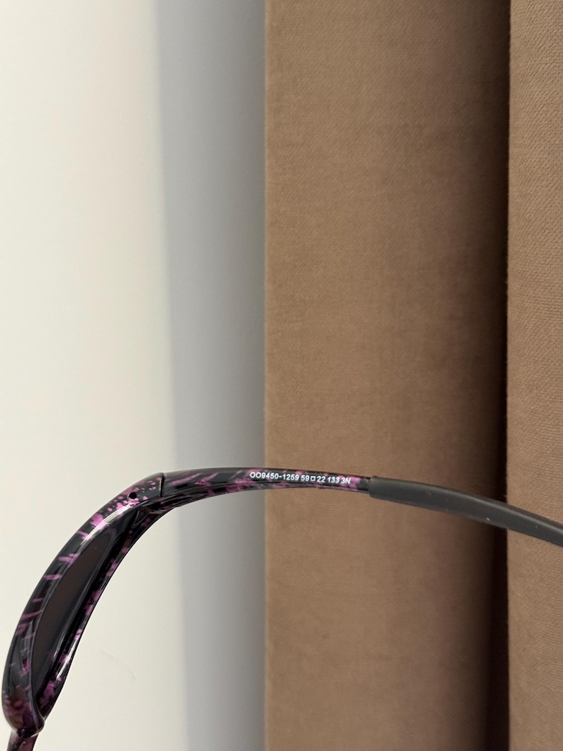 May include: Black sunglasses with a purple and black patterned frame. The glasses have a curved design and a dark lens. The text "008450-1259 59□22 135 3N" is printed on the frame.