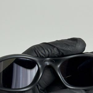 May include: Black sunglasses with a rounded frame and dark lenses. The sunglasses have the word "MINUTE" printed on the top of the frame. The sunglasses are held by a person wearing black gloves.